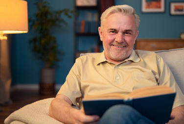 Older Man Reading A Book At Home Sat On The Sofa